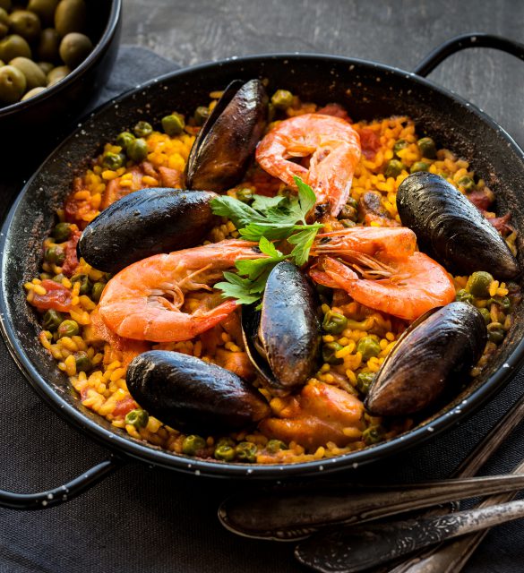 Paella in black pan with rice, shrimps, mussels, squid and meat, bowl with olives and vintage cutlery. Seafood paella, traditional spanish dish. Paella on rustic black wooden table. Selective focus